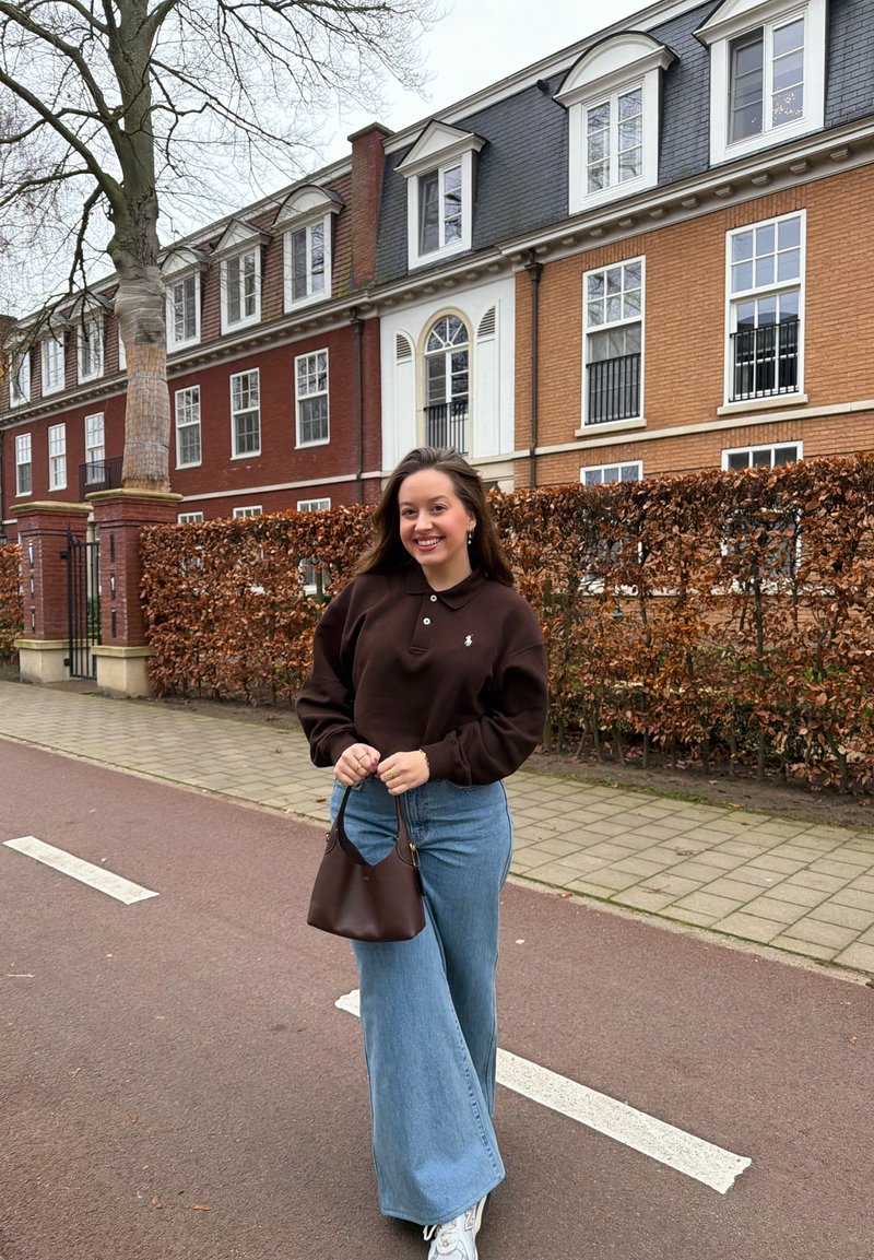 Jeune femme souriante tenant un sac à main marron, debout dans une rue avec des bâtiments en briques et des arbres sans feuilles en arrière-plan.