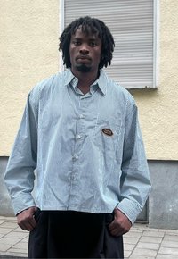 Camicia a righe in azzurro chiaro e bianco, con tasca frontale e toppa. Vestibilità ampia, maniche lunghe e colletto classico.