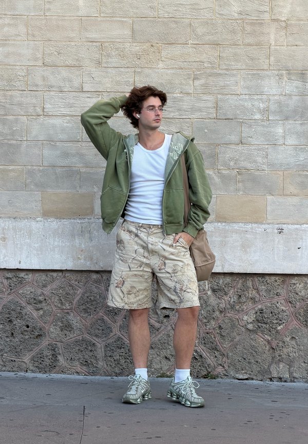 Jeune homme portant des lunettes, un sweat à capuche vert, un débardeur blanc, un short à motifs et des baskets, debout contre un mur en pierre, tenant un sac beige et touchant ses cheveux.
