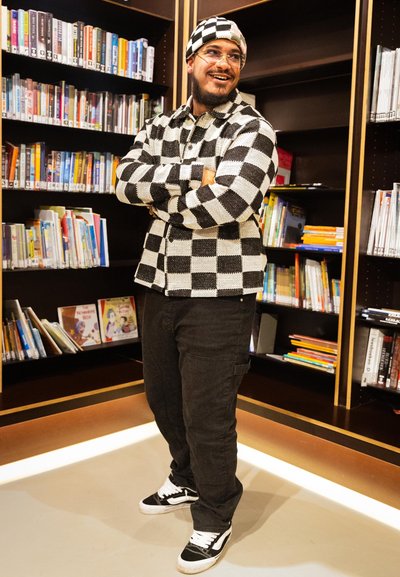 Chaqueta de punto a cuadros en blanco y negro, pantalones negros y zapatillas en blanco y negro. De pie frente a una estantería llena de libros.