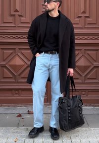 Černý kabát, černý svetr, světlé modré džíny, černé boty. Nese černou tašku s rukojetí. Stojí před texturovaným hnědým pozadím.