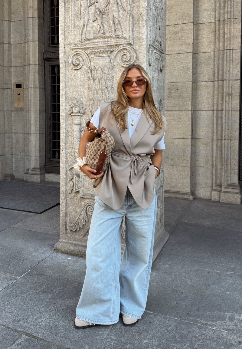 Gilet beige senza maniche sopra una t-shirt bianca, jeans larghi blu chiaro, che tiene una borsa a tracolla marrone con motivo, accessorata con collane a più strati.
