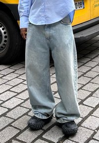 Lys blå oversized denimbukser med løs passform, med en falmet tekstur. Brukt med svarte teksturerte sko, plassert mot en brosteinsbelagt bakgrunn.