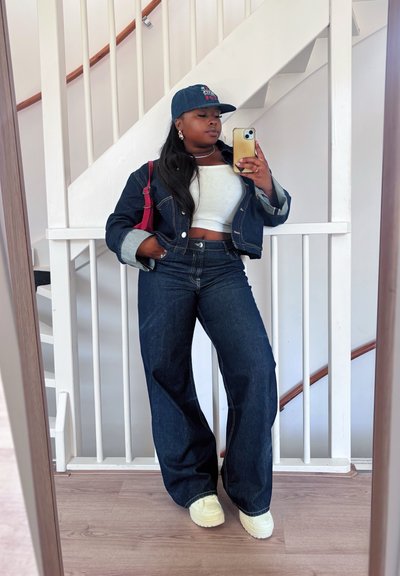 Chaqueta de mezclilla azul con puños en contraste, top corto blanco, jeans anchos azules, zapatos de plataforma amarillos y una gorra azul. Detalle de correa roja en el bolso.