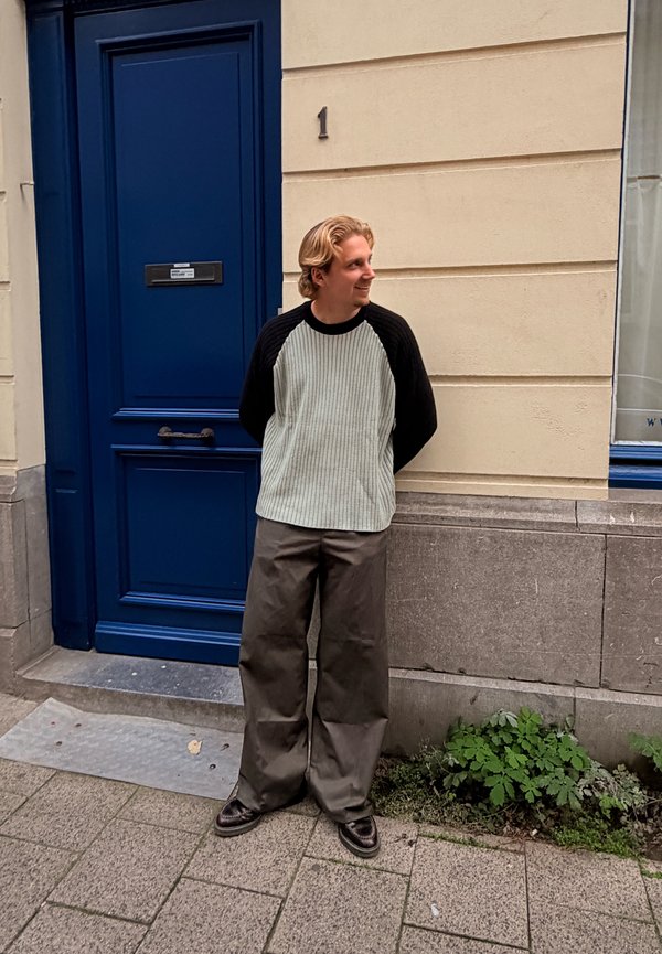 Lässiges Outfit mit einem gerippten, hellgrünen Pullover mit schwarzen Ärmeln und weiten, dunkelgrauen Hosen, vor einer blauen Tür und einer beigen Wand.