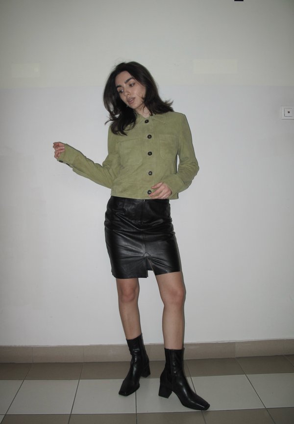Žena stojící uvnitř, oblečená v olivově zelené zapínané bundě, černé kožené sukni s rozparkem a černých kotníkových botách na podpatku na dlažbě.