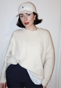 Pull en maille surdimensionné couleur crème avec poignets côtelés, porté par-dessus une chemise blanche. Casquette blanche avec logo, pantalon foncé et cheveux noirs courts.