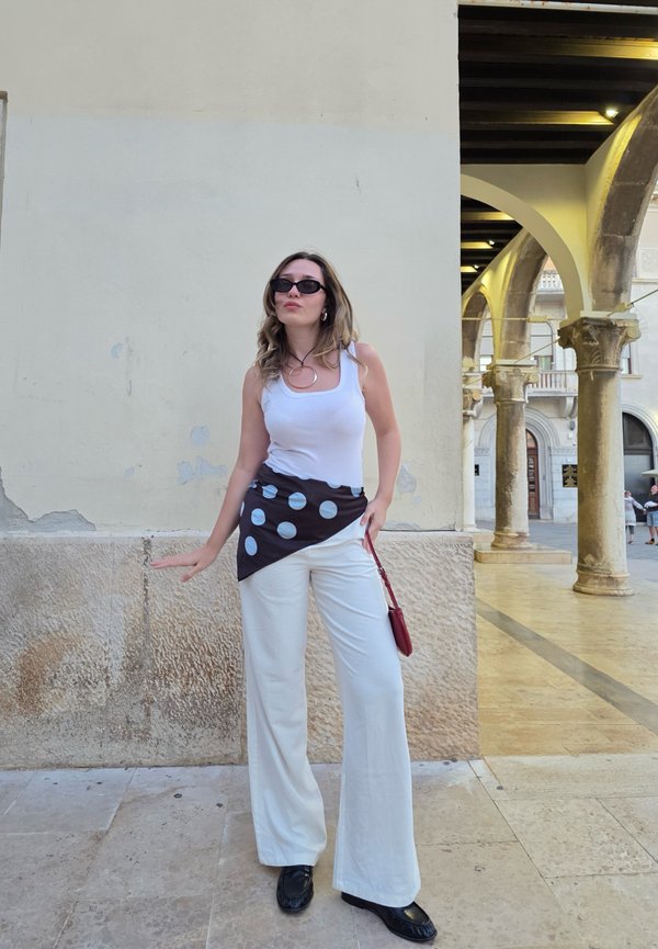 Débardeur blanc, étole à pois noir et blanc, pantalons larges blancs, chaussures noires et un sac à main rouge devant un mur en pierres.