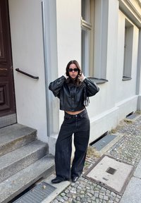 Donna con giacca di pelle nera, jeans neri a gamba larga e occhiali da sole, in piedi sul marciapiede di ciottoli accanto a un edificio bianco con scale.