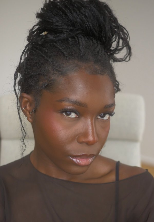 Jeune femme aux cheveux foncés tressés et attachés, portant un haut noir transparent, regardant directement la caméra avec une expression neutre.