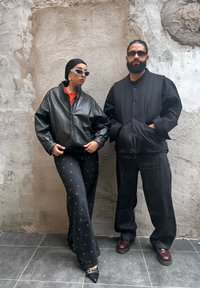 Schwarze Lederbomberjacke, orange Oberteil, schwarze gemusterte weite Hosen und spitze Absätze. Schwarze gepolsterte Weste und dunkle Jeans. Graue strukturierte Wand im Hintergrund.