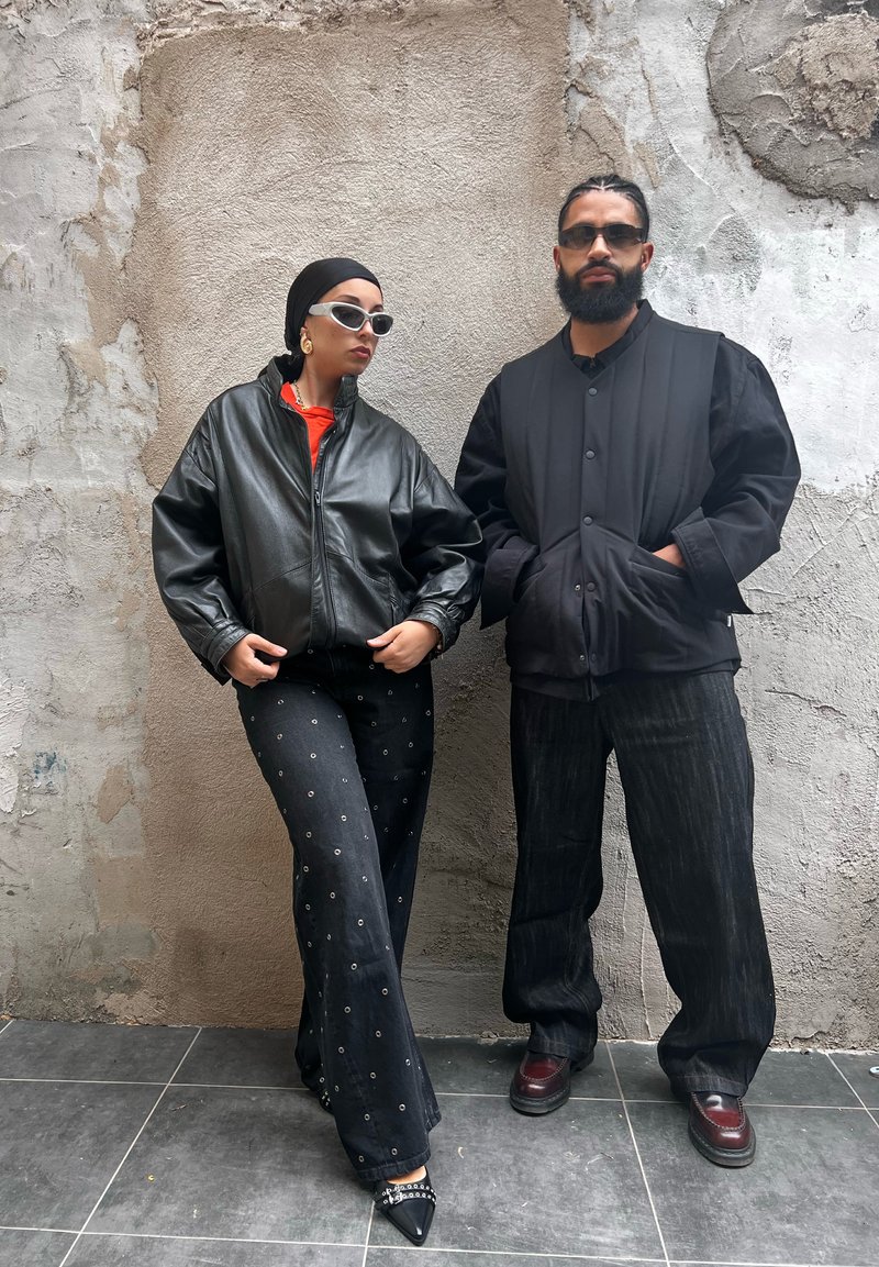Casaco bomber de couro preto, blusa laranja, calças largas pretas com padrão e sapatos de bico fino. Colete acolchoado preto e jeans escuros. Fundo de parede texturizada cinza.