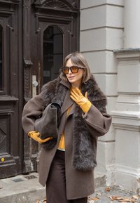 Barna gyapjúkabát szőrmekalappal, sárga pulóver, barna nadrág �és mustársárga kesztyűk. Szürke bőrtáskát tart. Nagy napszemüveg.