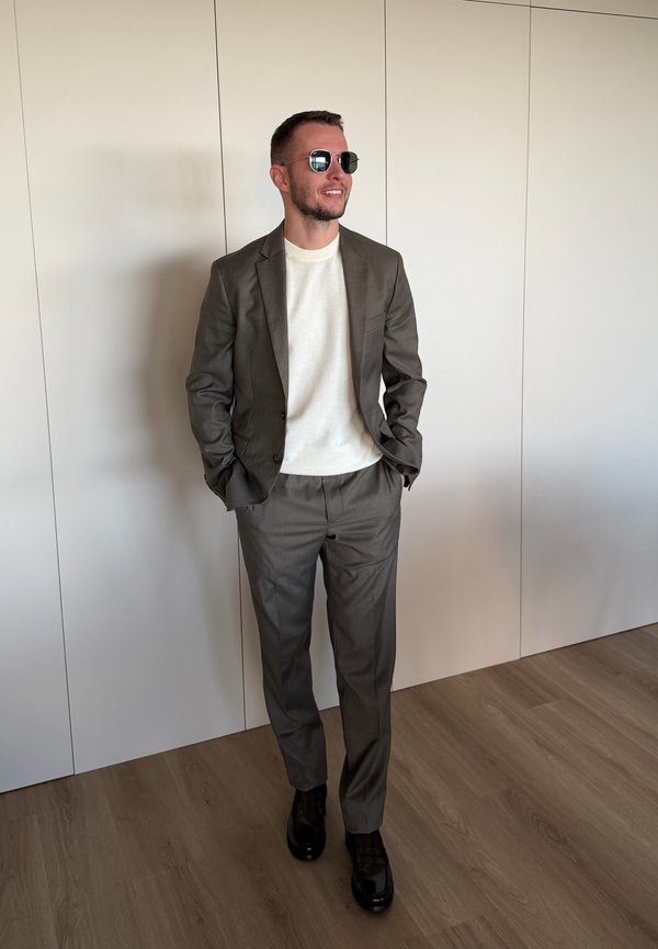 Hombre vestido con traje marrón, camisa blanca, zapatos negros y gafas de sol, de pie en el interior frente a una pared blanca con las manos en los bolsillos.