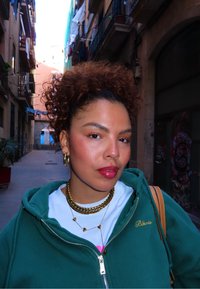 Grønn hettegenser med glidelås og broderi, hvit T-skjorte, lag på lag av gylne halskjeder, gullringøreringer og middels langt krøllete hår. Urban bakgrunn.