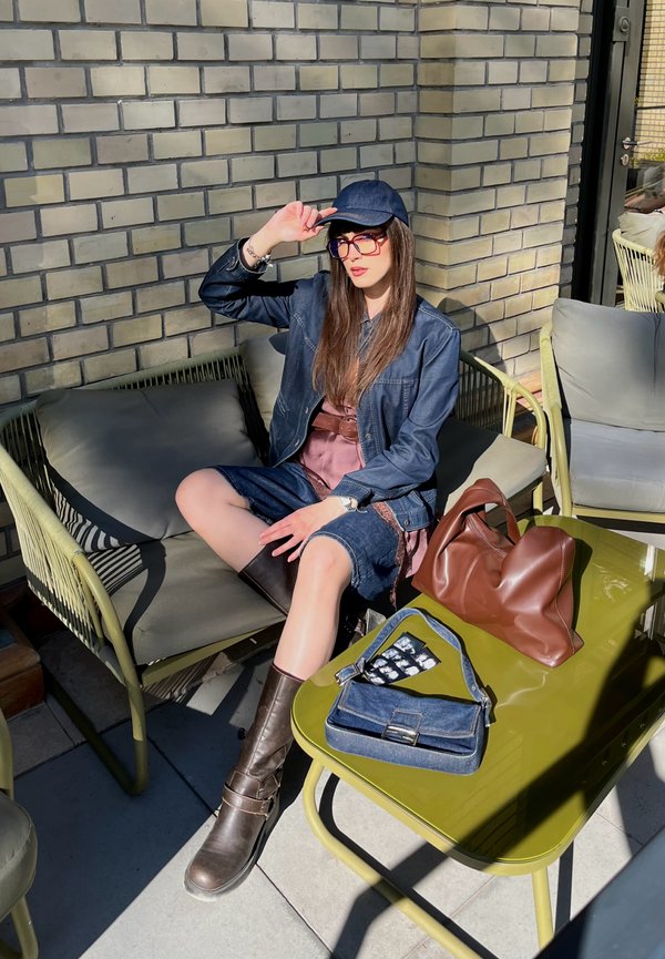 Vrouw in spijkerjasje, korte broek, laarzen en pet zittend op een buitensofa bij een olijftafel met twee handtassen in het zonlicht.