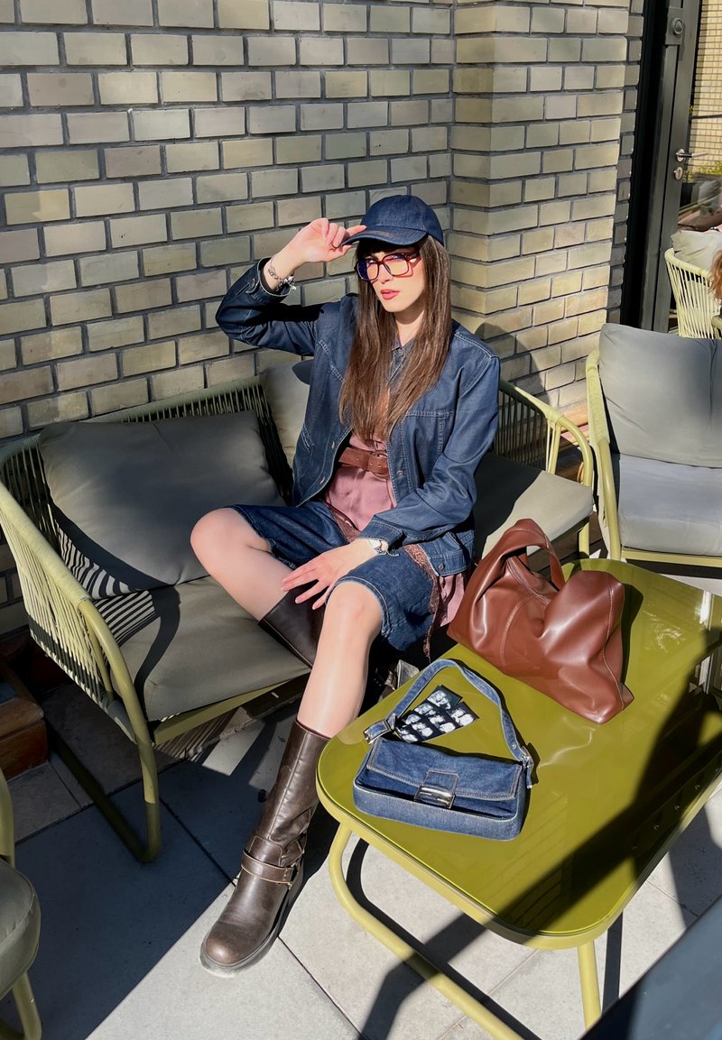 Femme en veste en jean, short, bottes et casquette assise sur un canapé d'extérieur près d'une table en olivier avec deux sacs à main en plein soleil.