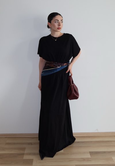 Mujer con el pelo oscuro recogido en un moño, vistiendo un vestido negro, cinturón de pañuelo estampado, sosteniendo un bolso burdeos, de pie contra una pared lisa.