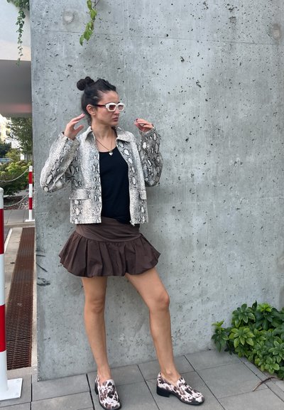 Chaqueta corta con estampado de serpiente, blusa negra, falda de volantes marrón y zapatos de lona estampados, todo frente a una pared de concreto y vegetación.