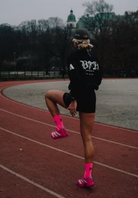 Fekete logós pulóvert és rövidnadrágot viselő sportoló élénk rózsaszín zoknival és cipővel fut a piros futópályán, háttérben fák.