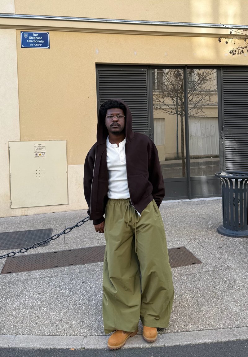 Άνδρας με γυαλιά, καφέ φούτερ με κουκούλα, λευκό πουκάμισο, φαρδιά πράσινα παντελόνια και μπεζ μποτάκια στέκεται στο δρόμο κοντά σε κτίριο με γυάλινες πόρτες και κάδο απορριμμάτων.