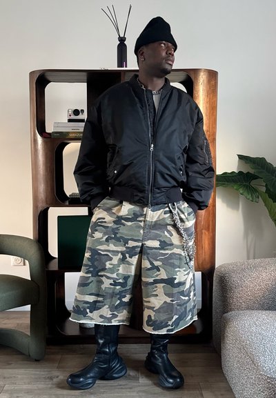 Hombre con gorro negro, chaqueta bomber negra, pantalones cortos anchos de camuflaje y botas negras, de pie en el interior cerca de una estantería y una planta.