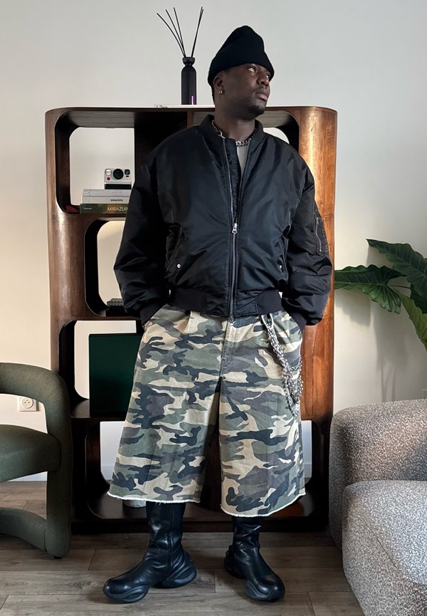 Man in zwarte muts, zwarte bomberjas, camouflage wijde shorts en zwarte laarzen, staand binnen bij een plank en een plant.