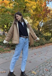Braune, mit Schafsfell gefütterte Jacke, schwarzes Polohemd, blaue Jeans und braune Stiefeletten. Steht im Freien mit Herbstlaub im Hintergrund.