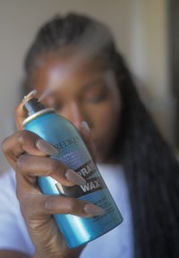 Spray Wax de Redken en un aerosol cilíndrico de color azul, etiquetado en blanco y negro. La boquilla está apuntando hacia arriba y se puede ver una fina niebla.