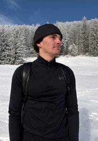 Jeune homme vêtu de vêtements d'hiver noirs et d'un bonnet, debout dans un paysage enneigé avec des arbres couverts de givre sous un ciel bleu.