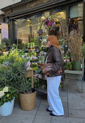 Mujer con pelo rojo, gafas de sol, chaqueta marrón y pantalones blancos está junto a macetas fuera de una floristería con varias plantas y flores.