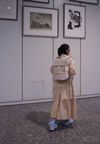 Mochila beige con una solapa y un bolsillo frontal, llevada con un abrigo trench beige claro y jeans azules, frente a un fondo de obras de arte enmarcadas.