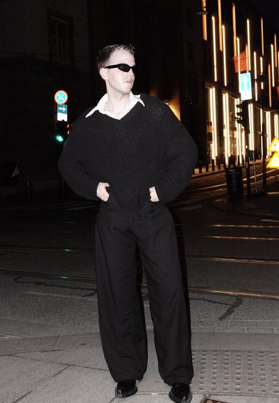 Suéter negro de punto oversize, camisa blanca con cuello debajo, pantalones negros holgados, gafas de sol negras, de pie en una calle por la noche.