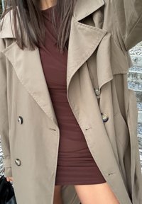 Trench-coat beige avec grands revers et deux boutons, porté sur une robe bourgogne ajustée. Tissu lisse, longueur genoux, détails minimaux.