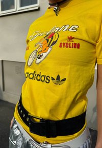 Žlté bavlnené tričko s ilustráciou cyklistu a logom "adidas". Kombinované s čiernym opaskom a striebornými metalickými nohavicami.