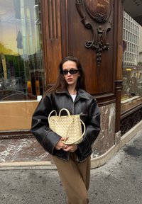 Fekete bőrből készült túlméretezett dzseki, krémszínű szövött kézitáska, barna nadrág. A háttérben fa és üveg bolti elemek láthatók.