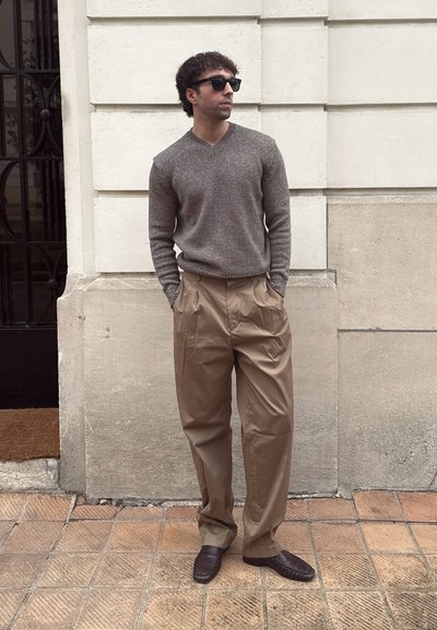 Hombre con gafas de sol, suéter gris, pantalones beige holgados y zapatos oscuros, de pie con las manos en los bolsillos contra una pared de piedra.