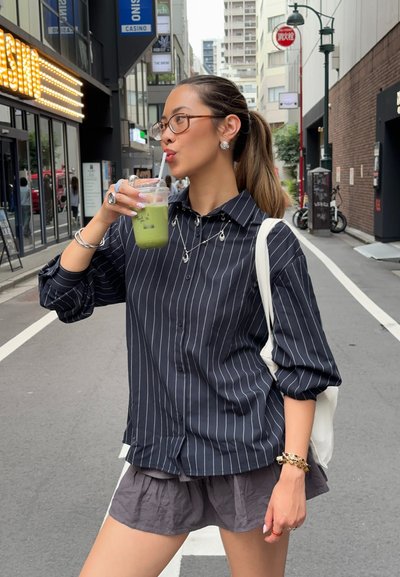 Camisa de botones a rayas blancas y negras, combinada con una falda gris, sosteniendo una bebida verde en un vaso transparente, joyería de plata acentuando el atuendo.