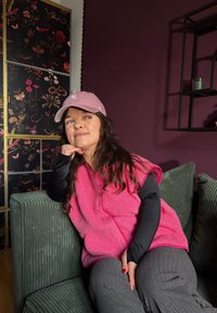 Jeune femme portant une casquette et un gilet roses, assise sur un canapé vert, reposant son menton sur sa main, avec une œuvre d'art florale sombre et un mur violet derrière elle.