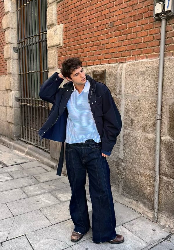 Jachetă supradimensionată de culoare bleumarin închis peste un tricou polo cu dungi albastre deschise, asociată cu blugi largi din denim închis și pantofi maronii. Fundal cu zid de cărămidă.
