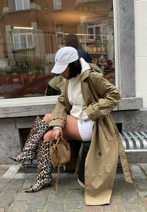 Trinchera beige sobre una sudadera blanca, combinada con botas hasta la rodilla de estampado de leopardo y un pequeño bolso marrón. Fondo de concreto.