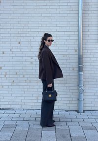 Γυναίκα με μακριά κυματιστά μαλλιά που φοράει γυαλιά ηλίου, σκούρο σακάκι, φαρδιά παντελόνια, κρατώντας μια μαύρη τσάντα, στέκεται μπροστά σε έναν λευκό τοίχο από τούβλα με ένα μεταλλικό σωλήνα.