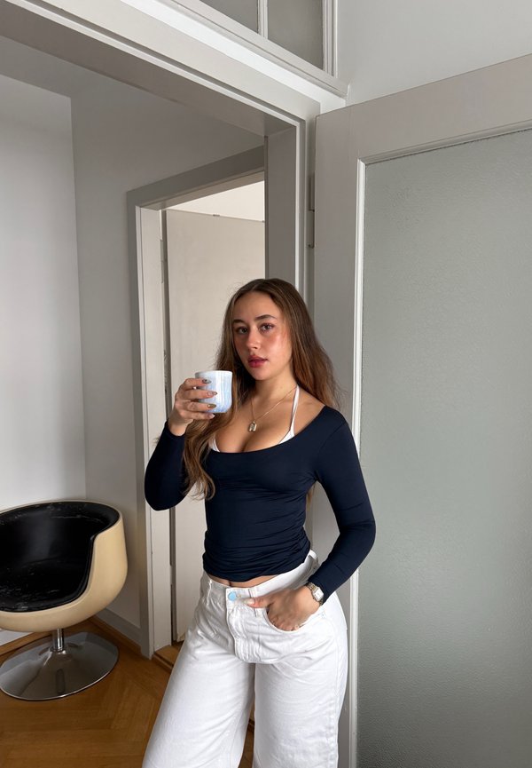 Femme aux longs cheveux portant un haut noir et un pantalon blanc, tenant une tasse mouchetée, debout dans une porte à côté d'une chaise moderne noire et beige.