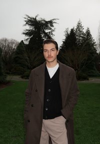 Manteau marron à double boutonnage porté sur un cardigan noir et un t-shirt blanc, associé à un pantalon beige clair. Herbe verte et arbres en arrière-plan.