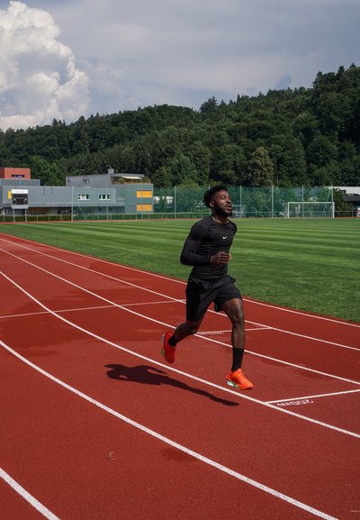 Corredor atlético negro con una camiseta negra de manga larga ajustada y pantalones cortos, zapatos de correr naranjas, corriendo a toda velocidad en una pista roja con hierba verde al fondo.