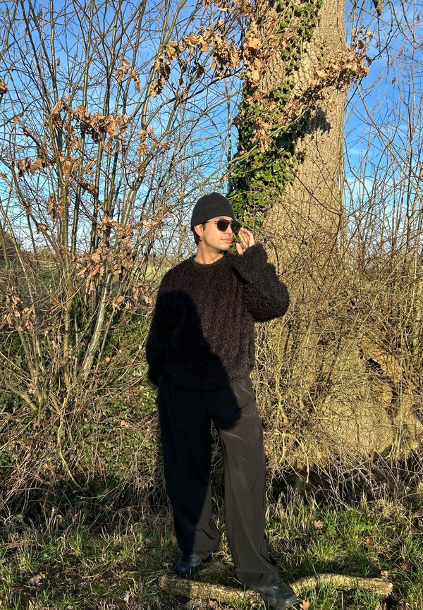 Un pull noir en peluche, un pantalon noir ample, un bonnet en tricot et des lunettes de soleil. Cadre naturel avec des arbres dépouillés et un ciel bleu clair.