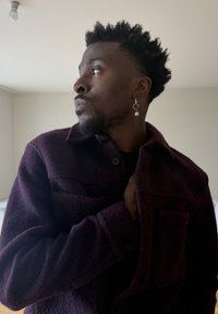 Homme aux cheveux courts et bouclés portant une boucle d'oreille en perle et une veste boutonnée violet foncé, regardant vers la gauche dans une pièce minimaliste.