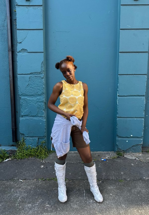 Nuori henkilö, jolla on kaksi afro-puff-kampausta, seisoo sinistä seinää vasten, päällään keltainen hihaton toppi, ruskeat shortsit, valkoiset saappaat ja harmaa collegepaita vyötäröllä sidottuna.