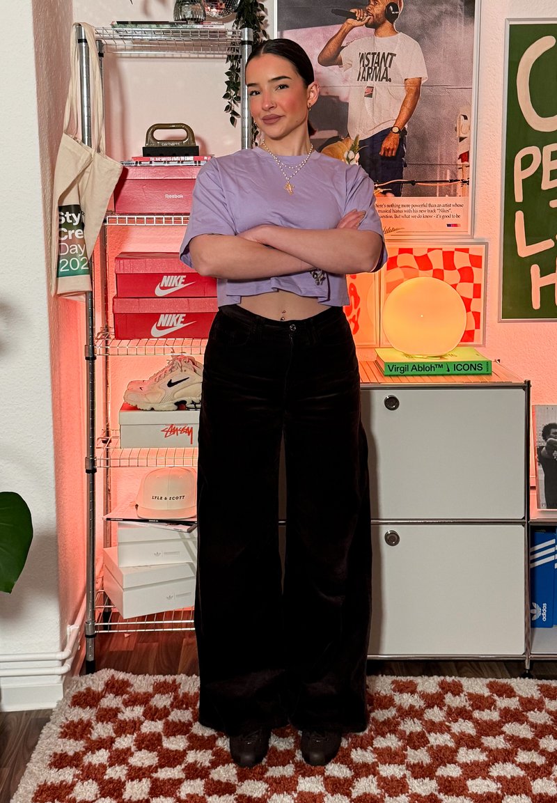 Jeune femme portant un haut violet court et un pantalon noir, debout bras croisés dans une pièce avec des boîtes de baskets, des affiches et une lampe ronde lumineuse.