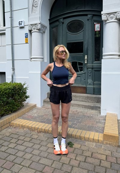 Shorts atléticos negros y una camiseta corta ajustada de color azul marino, combinados con zapatillas blancas y naranjas. De pie sobre un camino de piedra texturizado cerca de una puerta verde.